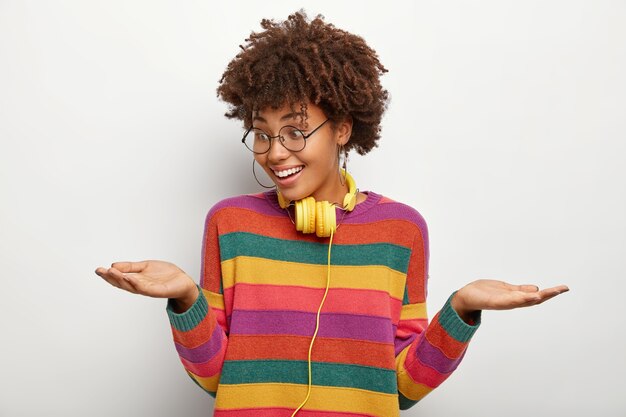 Positive woman raises palms, holds two invisible objects, makes decision between good proposals, wears optical glasses and striped sweater