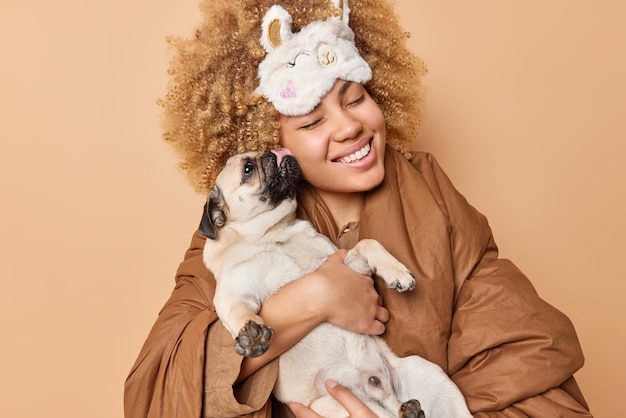 ポジティブな女性は、パグ犬が家畜と一緒に寝る毛布に包まれたsleepmaskを身に着けているお気に入りのペットのポーズからキスを受け取ります家でのんびりとした一日を過ごします友情の概念