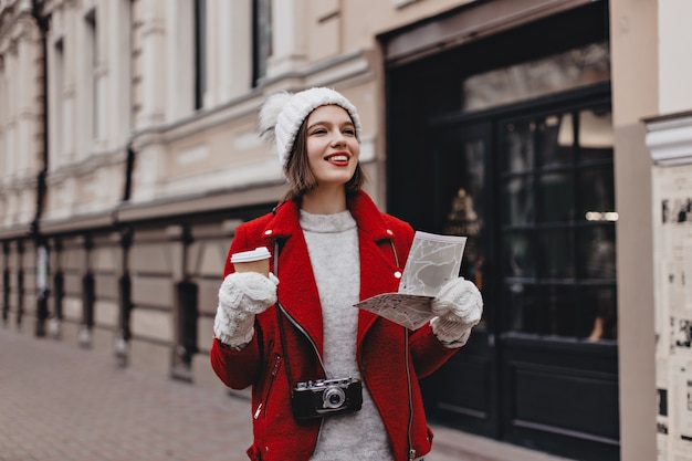 Позитивная женщина в красной теплой куртке, кашемировом свитере и белой шляпе с перчатками гуляет по городу с кофе. Туристка с ретро камерой на шее держит карту.