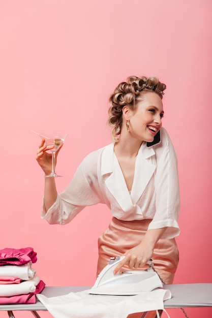 Positive woman in silk blouse with smile talking on phone, ironing clothes and holding martini glass