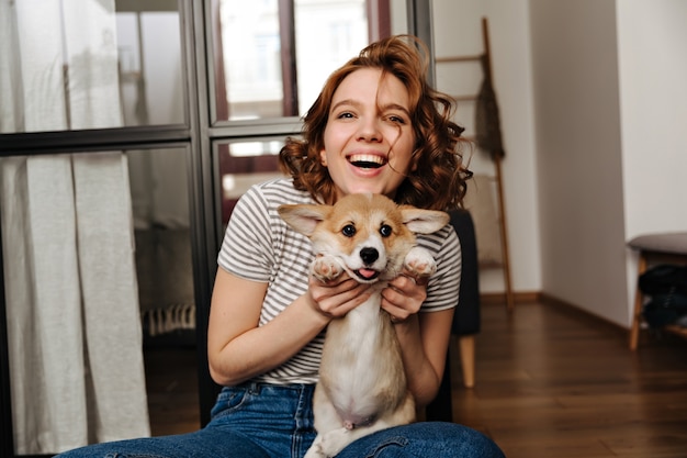 Positive woman sits on floor in living room and with smile plays with her beloved dog.