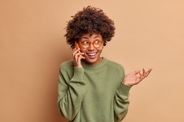 Positive woman smiles broadly looks aside keeps hand raised giggles happily has funny conversation wears optical glasses and jumper isolated over brown wall