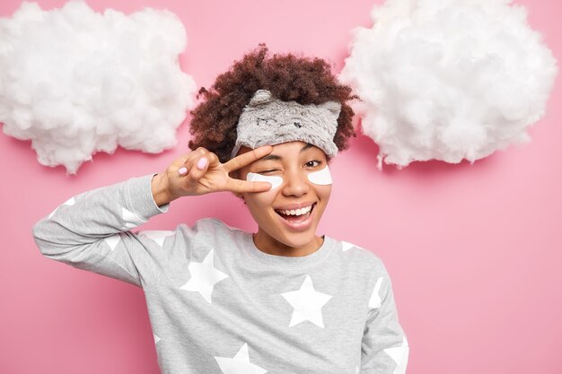 Positive woman smiles gladfully makes peace gesture over eye winks eyes applies collagen patches under eyes dressed in pajama isolated over pink wall white clouds above