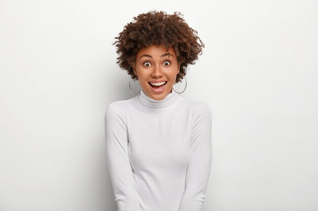 Positive woman smiles pleasantly, wears silver earrings and casual poloneck sweater