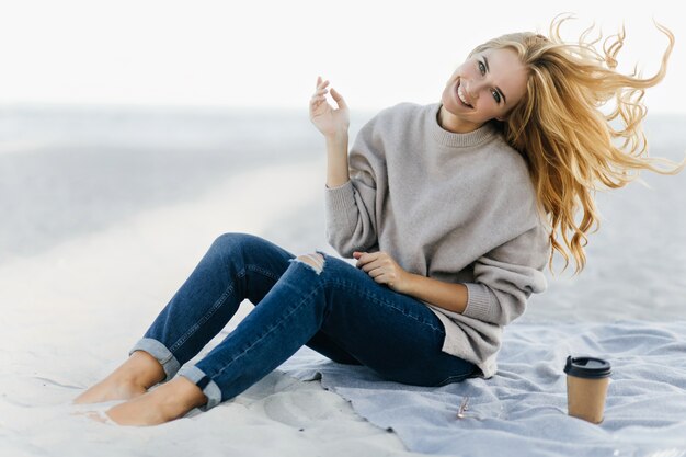 Позитивная женщина в мягком свитере дурачится на пляже. Открытый портрет очаровательной женской модели, сидящей в песке с чашкой чая.