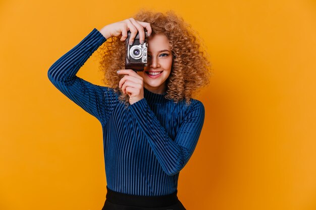 Positive woman in striped blue sweater takes photo on retro camera on yellow space.
