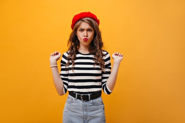 Positive woman in striped shirt and beret makes funny face. Curly young lady in denim skirt with wide black belt posing on isolated background.