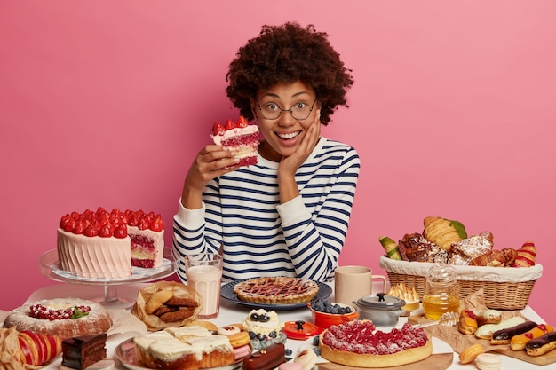 La donna positiva golosa di dolci assaggia una deliziosa torta di fragole, interrompe la dieta e mangia cibo con molte calorie, si siede a un grande tavolo con dolciumi