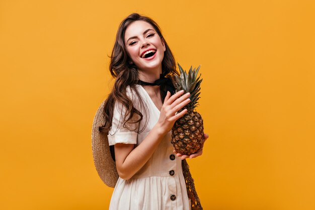 Positive woman in white sundress and straw hat laughs while holding pineapple.