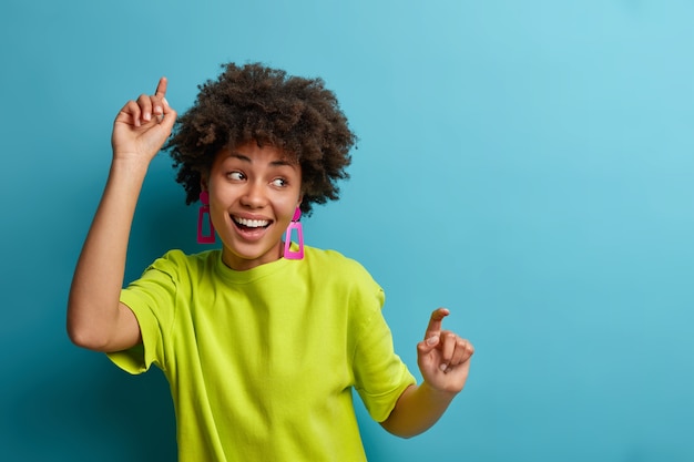 Positive woman with Afro hairstyle dances with arms raised, feels carefree and upbeat, extremly happy and expresses joy, wears green t shirt, isolated over blue wall, moves energetically