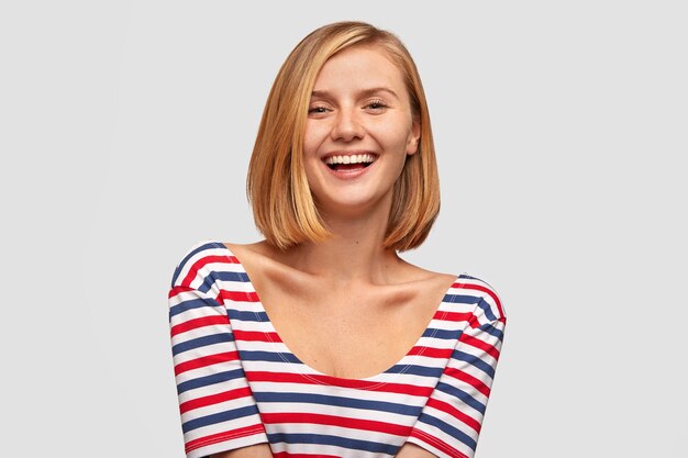 Positive woman with broad smile, shows white teeth, laughs at good joke, likes funny story from interlocutor, has slim body, dressed in striped jacket