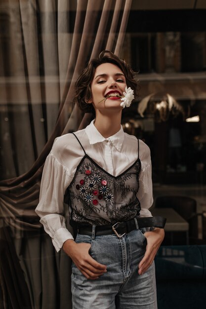 Positive woman with brunette hair in blouse with lace and jeans poses with flower in mouth inside. Wavy haired woman with red lips smiles in cafe.