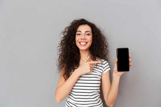 灰色の壁に対してポーズをとって彼女のスマートフォンを広告するような人差し指を指している白人の外観を持つ肯定的な女性