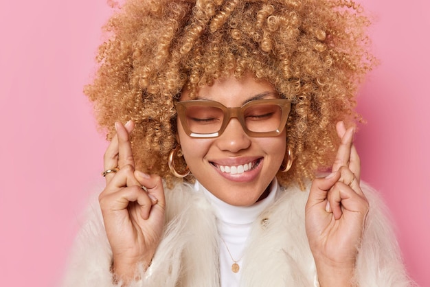 Free photo positive woman with cheerful expression smiles broadly keeps fingers crossed believes in good luck wears winter coat isolated over pink background anticipates something pleads feels hopeful.