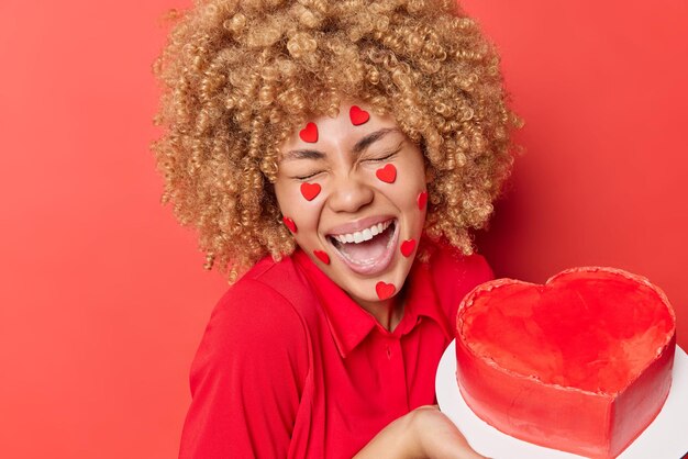 巻き毛のブロンドの髪のポジティブな女性は喜んで目を閉じて笑う誕生日を祝うか、お祝いのイベントは心で飾られた顔が赤い背景の上に分離された甘いおいしいケーキを保持しています