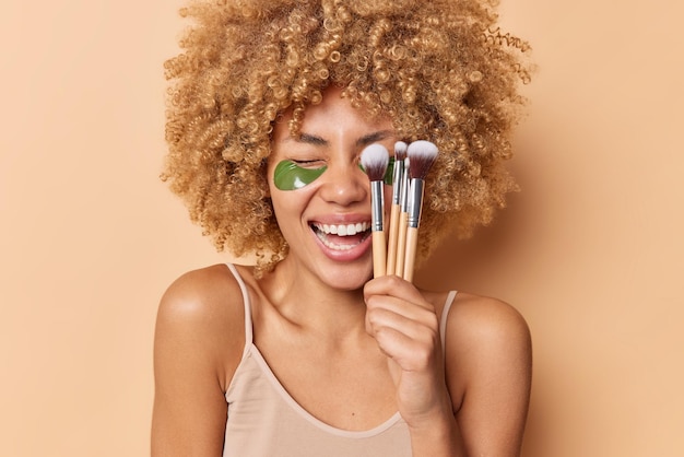 Positive woman with curly bushy hair holds cosmetic brushes over face giggles happily applies green hydrogel patches under eyes for skin treatment wears casual t shirt isolated over beige studio wall