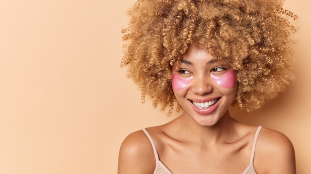 곱슬곱슬한 머리를 가진 긍정적인 여성은 행복한 표정을 짓고 있으며 건강한 깨끗한 피부를 가지고 있으며 잘 관리된 안색은 눈 아래에 분홍색 하이드로겔 패치를 적용합니다.