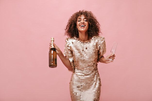 Positive woman with curly fluffy hairstyle in shiny modern clothes laughing and holding bottle with wine and glass on pink wall..