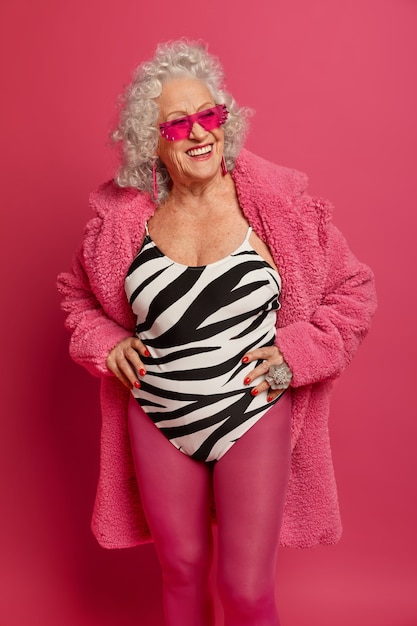 Positive woman with gray hair, smiles broadly, keeps hands on waist, keeps fit and healthy, wears trendy sunglasses, swimsuit, pink tights and robe, dreams about vacation during self isolation