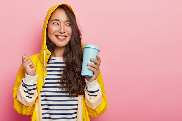 Positive woman with long dark straight hair, raises clenched fist, holds takeaway coffee, dressed in striped jumper, yellow raincoat