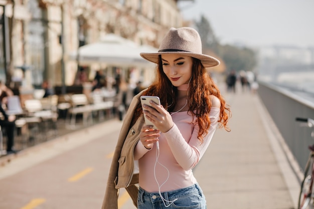 Foto gratuita donna positiva con acconciatura lunga utilizza lo smartphone mentre si sta in piedi sull'argine