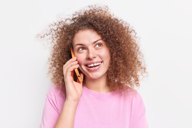 Positive woman with natural curly hair calls friend to meet up holds smartphone near ear looks upwards happily enjoys funny conversation wears casual pink t shirt isolated over white wall