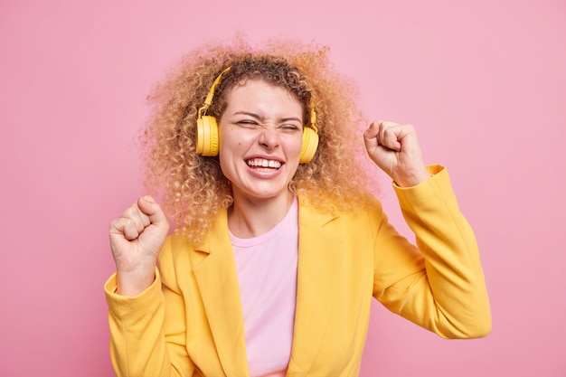 Positive woman with natural curly hair dances carefree makes triumph dance smiles broadly enjoys listening audio track via wireless headphones 