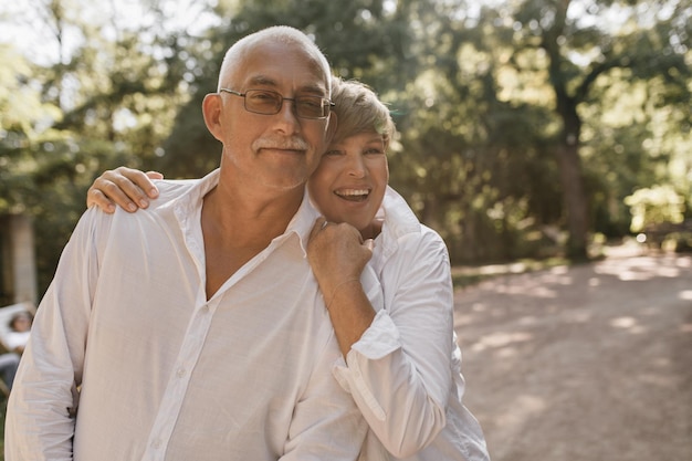 Positive woman with short hairstyle laughing and hugging grey haired man with mustache in cool eyeglasses and white shirt outdoor