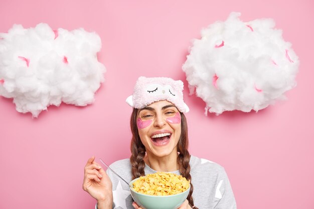 Positive woman with two pigtails smiles broadly wears slumber suit and sleepmask eats cereals for breakfast isolated over pink wall