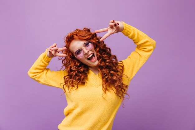 Positive woman in yellow sweater and glasses shows peace sign and laughs on lilac wall