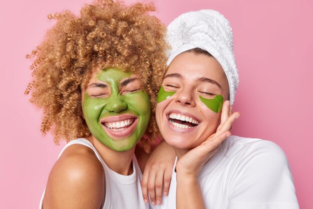 Positive women feel joyful smile broadly keep eyes closed apply green nourishing mask and patches dressed in white t shirts undergo beauty procedures isolated over pink background Facial treatment