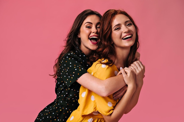Free Photo positive women in polka dot dresses hug on pink background charming girls in a good mood in summer clothes are smiling at camera