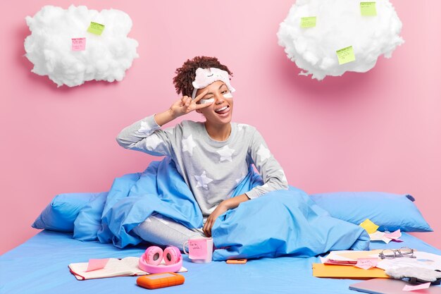 Positive young African American woman makes peace gesture sticks out tongue enjoys domestic atmosphere learns material from home 