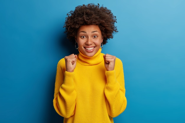 Positive young Afro American woman smiles broadly and wears yellow jumper isolated over blue background.