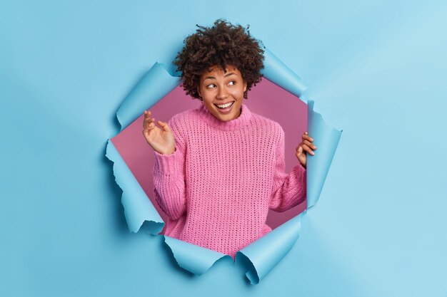 Positive young Afro American woman smiles happily keeps hand raised has carefree expression