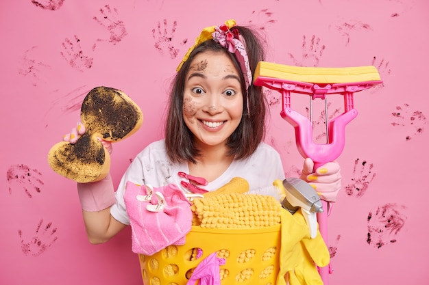 Positive young Asian woman holds dirty sponge and mop busy doing home daily routine washes laundry removes dirt everywhere isolated over pink studio wall