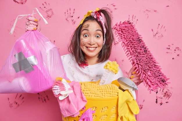 Positive young Asian woman picks litter in polythene bag holds dirty mop cares about purity has dirty face busy doing laundering isolated on pink wall