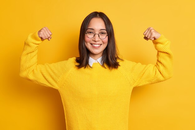 Позитивная молодая азиатка поднимает руки, демонстрирует мускулы, притворяется очень сильными и мощно улыбается, нежно носит повседневный свитер.