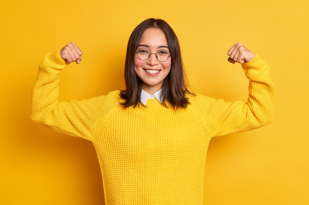 Бесплатное фото Позитивная молодая азиатка поднимает руки, демонстрирует мускулы, притворяется очень сильными и мощно улыбается, нежно носит повседневный свитер.