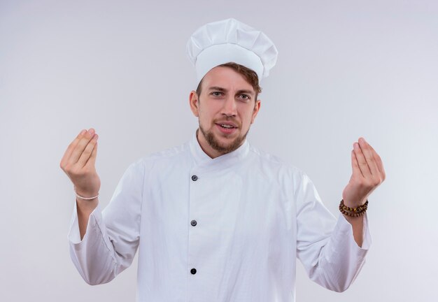 A positive young bearded chef man wearing white cooker uniform and hat holding fingers together hand gesture while looking on a white wall