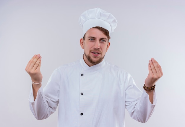Позитивный молодой бородатый шеф-повар в белой униформе и шляпе, держащий пальцы вместе, жест рукой, глядя на белую стену