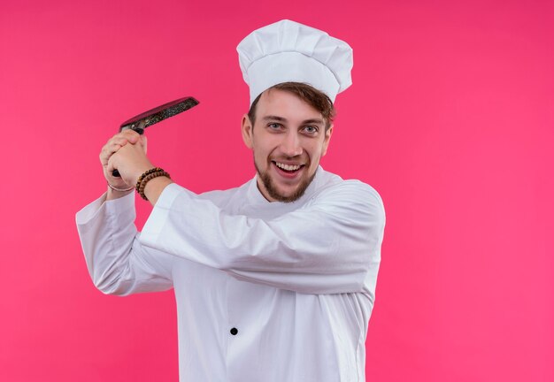 A positive young bearded chef man in white uniform holding frying pan like a baseball bat while looking on a pink wall