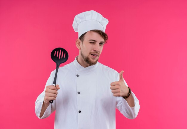 A positive young bearded chef man in white uniform holding slotted spoon and showing thumbs up while looking on a pink wall
