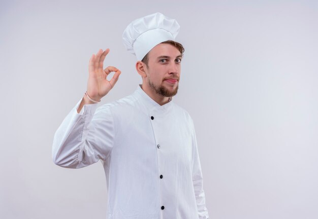 A positive young bearded chef man in white uniform showing ok gesture on a white wall