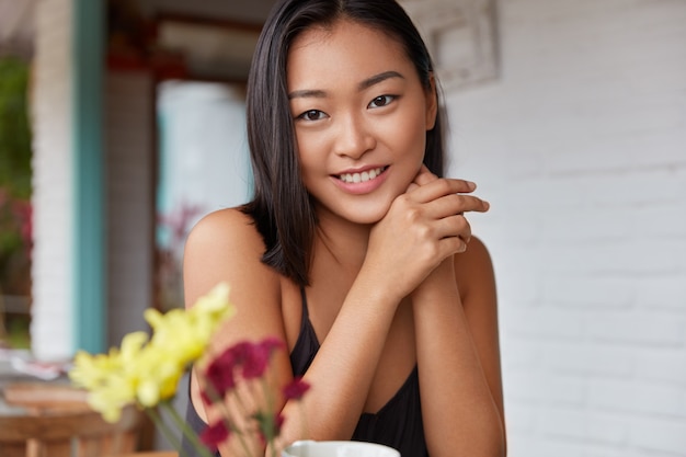Позитивная молодая красивая азиатка с широкой теплой улыбкой, с темными волосами и здоровой кожей, довольна полноценным отдыхом и обслуживанием в ресторане. Естественная красота