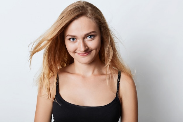 Positive young beautiful female with luxuriant blonde hair poses against white studio