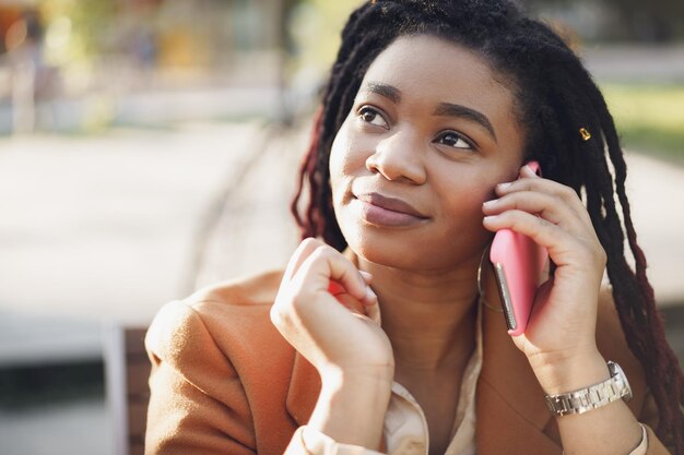 Позитивная молодая темнокожая женщина сидит в летнем кафе и разговаривает по телефону