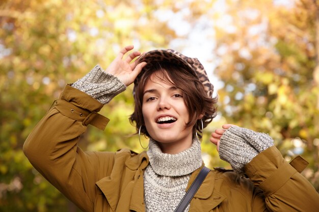 Positive young brown-eyed pretty brunette woman with bob hairstyle keeping her cap with raised hand while looking , wearing warm stylish clothes while walking through the park