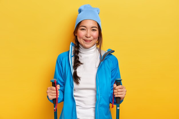 Positive young brunette woman enjoys nordic walking, holds trekking sticks, trains on forest trail, wears blue hat, jacket and white turtleneck, poses against yellow background. hiking and camping