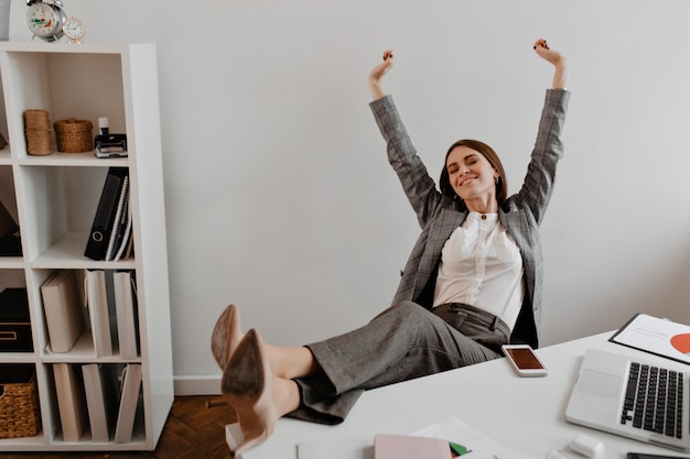 Free photo positive young business lady leans back in her chair and raises her arms up with satisfaction against shelves of documents.